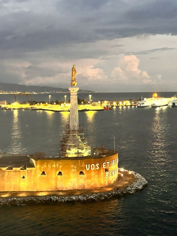 Estátua dourada iluminada sobre a coluna na entrada do porto de Messina, ao entardecer