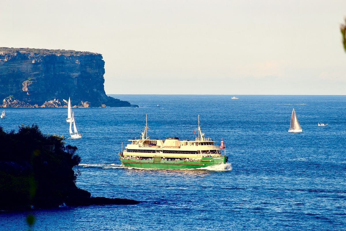 Ferry verde e amarelo de Manly atravessando o porto de Sydney, com veleiros e rochedos ao fundo